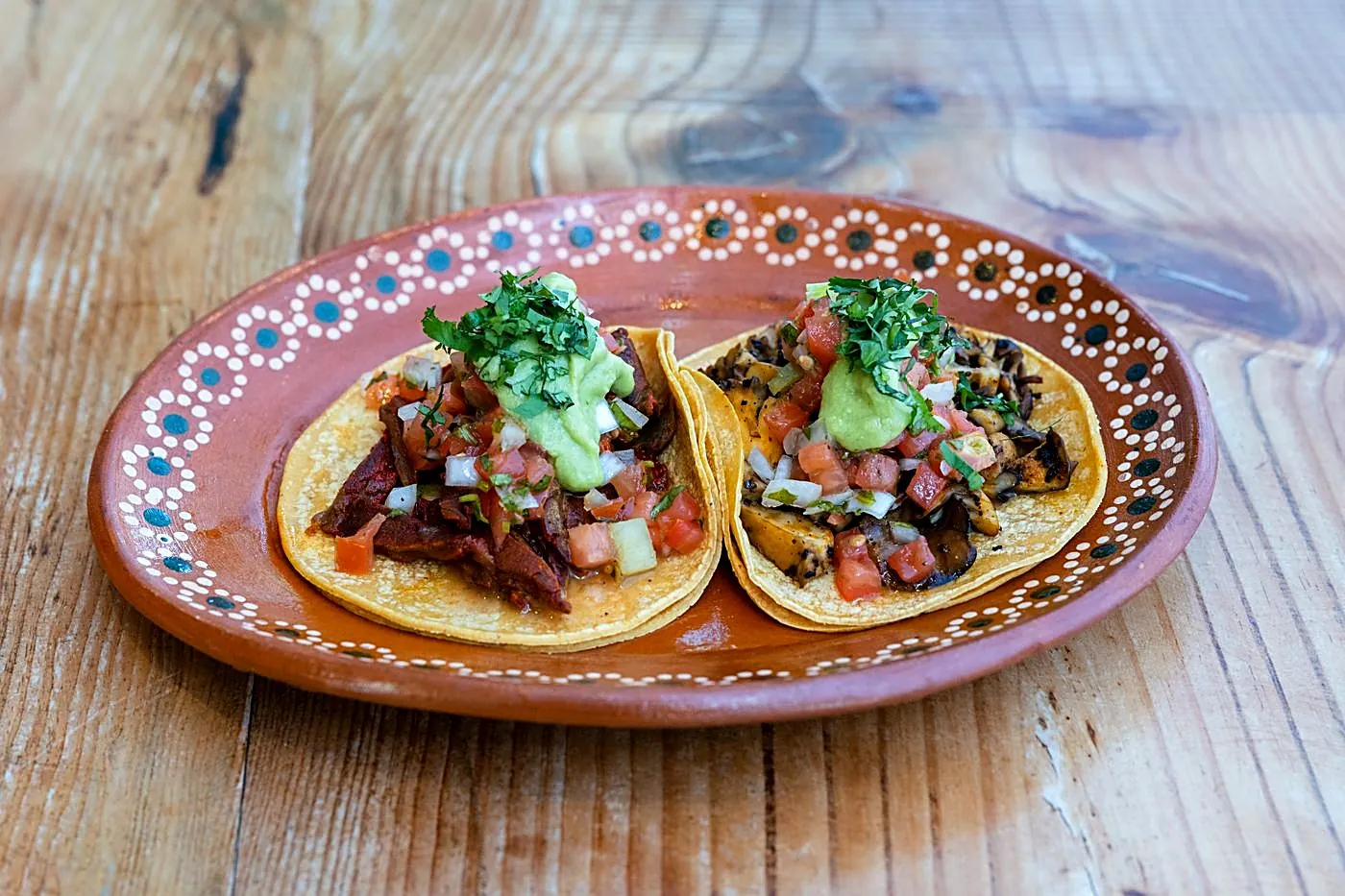 Two lengua (beef tongue) street tacos on a hand-painted clay plate, topped with pico de gallo and guacamole